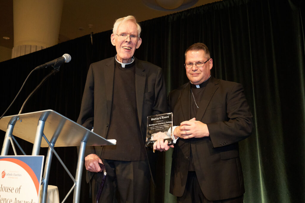 Monsignor Fahey smiles at the camera, holding his 2022 Bishop's Award from Catholic Charities. Bishop Lucia is standing next to him.