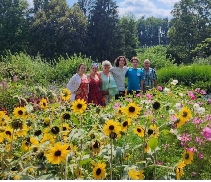 Tom and Lisa, with their four children, in a field of sunflowers. They have their arms around each other and they are smiling at the camera.