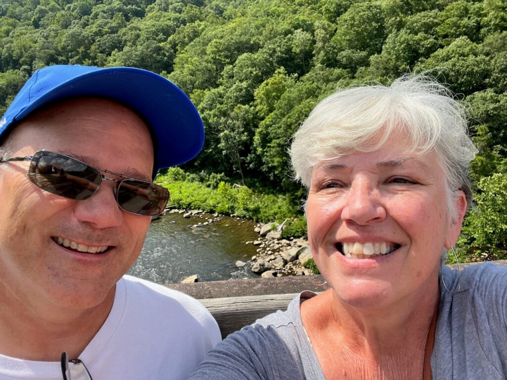 Tom and Lisa smile for the camera for a selfie - present day. They are by a body of water.